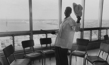 Movie still from “Salut les Cubains” (1963), directed by Agnès Varda – A black and white photo of a man holding a hat in front of a window; Medium shot, Low angle