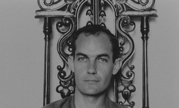 Movie still from “Salut les Cubains” (1963), directed by Agnès Varda – A black - and - white photo of a man in front of an ornate iron gate; Close Up shot, Overhead angle