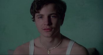 Movie still from “Salò, or the 120 Days of Sodom” (1975), directed by Pier Paolo Pasolini – A young man wearing a white tank top and a necklace; Close Up shot, Over the shoulder angle