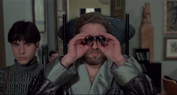 Movie still from “Salò, or the 120 Days of Sodom” (1975), directed by Pier Paolo Pasolini – A man with a beard and glasses looking through a pair of antennas; Medium shot, Over the shoulder angle