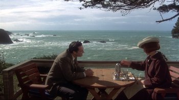 Movie still from “Same Time, Next Year” (1978), directed by Robert Mulligan – A man sitting at a wooden table near the ocean; Wide shot, High angle
