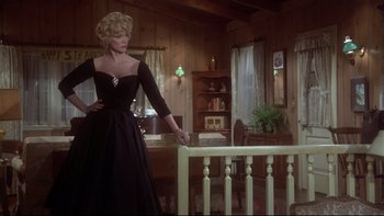 Movie still from “Same Time, Next Year” (1978), directed by Robert Mulligan – A woman in a black dress standing in front of a railing in a living room; Medium shot, Low angle