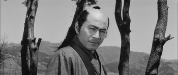 Movie still from “Samurai Rebellion” (1967), directed by Masaki Kobayashi – A man with long black hair is standing next to a tree; Close Up shot, Low angle