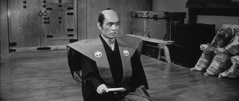 Movie still from “Samurai Rebellion” (1967), directed by Masaki Kobayashi – A man sitting on the ground wearing a robe and holding a book; Medium shot, Low angle