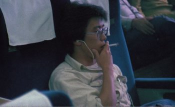 Movie still from “Sans Soleil” (1983), directed by Chris Marker – A man sitting on a plane smoking a cigarette and listening to music; Close Up shot, High angle