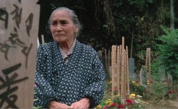 Movie still from “Sans Soleil” (1983), directed by Chris Marker – An older woman sitting in front of a flower garden; Medium shot, Low angle