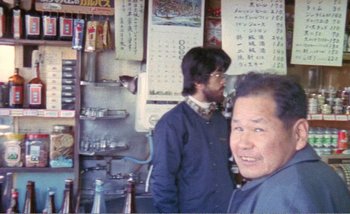 Movie still from “Sans Soleil” (1983), directed by Chris Marker – Two men standing next to each other in front of a menu; Medium shot, High angle