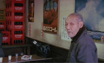 Movie still from “Sans Soleil” (1983), directed by Chris Marker – An older man sitting at a table in front of posters on the wall; Close Up shot, High angle
