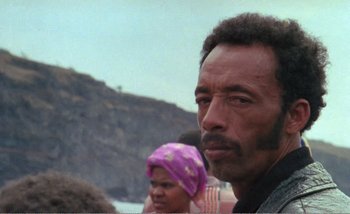 Movie still from “Sans Soleil” (1983), directed by Chris Marker – A man with a purple head scarf is standing in front of a group of people; Close Up shot, Over the shoulder angle