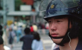 Movie still from “Sans Soleil” (1983), directed by Chris Marker – A man wearing a police helmet on a street; Close Up shot, High angle