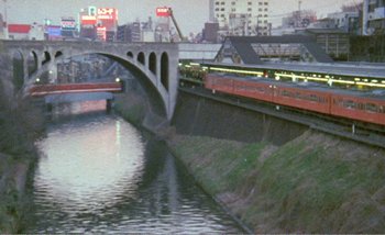 Movie still from “Sans Soleil” (1983), directed by Chris Marker – A train traveling over a bridge next to a river; Extreme Wide shot, High angle