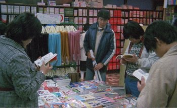 Movie still from “Sans Soleil” (1983), directed by Chris Marker – A group of people standing around a table with books on top of it; Medium shot, High angle