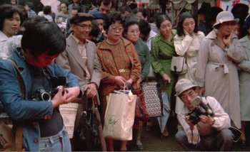 Movie still from “Sans Soleil” (1983), directed by Chris Marker – A group of people standing around each other holding shopping bags; Wide shot, High angle