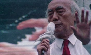 Movie still from “Sans Soleil” (1983), directed by Chris Marker – An older man holding a microphone up to his mouth; Close Up shot, Low angle