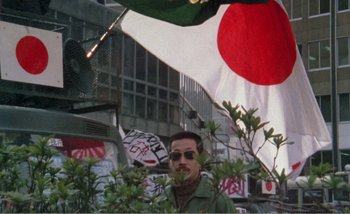 Movie still from “Sans Soleil” (1983), directed by Chris Marker – A man in a green jacket standing next to an asian flag; Medium shot, Low angle