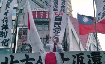 Movie still from “Sans Soleil” (1983), directed by Chris Marker – A man in a white shirt is speaking at a rally; Wide shot, High angle