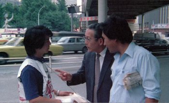 Movie still from “Sans Soleil” (1983), directed by Chris Marker – A man talking to two other men on the street; Medium shot, Over the shoulder angle