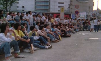 Movie still from “Sans Soleil” (1983), directed by Chris Marker – A group of people sitting on the side of the street; Extreme Wide shot, High angle