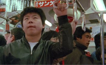 Movie still from “Sans Soleil” (1983), directed by Chris Marker – A man holding a pair of scissors in his hand while sitting on a bus; Medium shot, Low angle