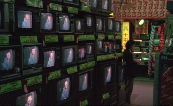Movie still from “Sans Soleil” (1983), directed by Chris Marker – A person standing in front of a bunch of televisions; Wide shot, High angle