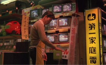 Movie still from “Sans Soleil” (1983), directed by Chris Marker – A man standing in front of a wall of tvs; Medium shot, Low angle