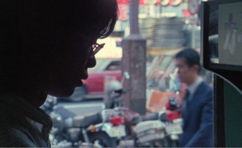 Movie still from “Sans Soleil” (1983), directed by Chris Marker – A man with glasses is looking at a man on a moped; Close Up shot, Over the shoulder angle