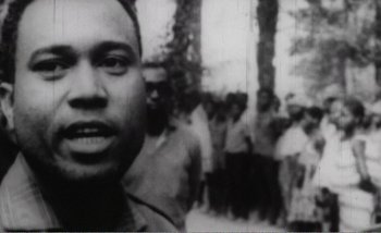 Movie still from “Sans Soleil” (1983), directed by Chris Marker – A black - and - white photo of a man in front of a group of people; Close Up shot, Low angle