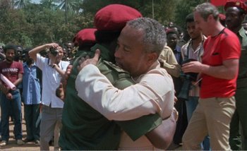 Movie still from “Sans Soleil” (1983), directed by Chris Marker – Two men hugging each other in front of a crowd of onlookers; Medium shot, Over the shoulder angle