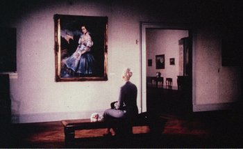 Movie still from “Sans Soleil” (1983), directed by Chris Marker – A woman sitting on top of a bench in front of a painting; Wide shot, High angle
