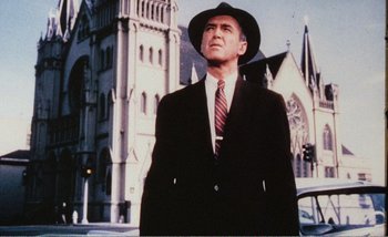 Movie still from “Sans Soleil” (1983), directed by Chris Marker – A man in a suit and hat standing in front of a church; Medium shot, Low angle