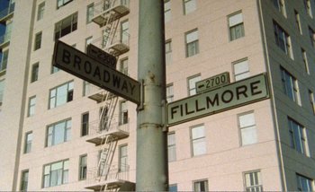 Movie still from “Sans Soleil” (1983), directed by Chris Marker – A pole with street signs on it in front of a tall building; Extreme Wide shot, High angle