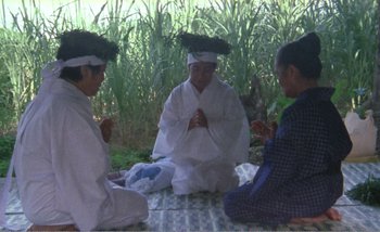 Movie still from “Sans Soleil” (1983), directed by Chris Marker – A group of people sitting on the ground in front of a forest; Wide shot, High angle