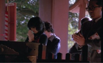 Movie still from “Sans Soleil” (1983), directed by Chris Marker – Two people are praying in front of a fence; Medium shot, Low angle