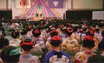 Movie still from “Sans Soleil” (1983), directed by Chris Marker – A group of people sitting in chairs in front of an audience; Extreme Wide shot, High angle