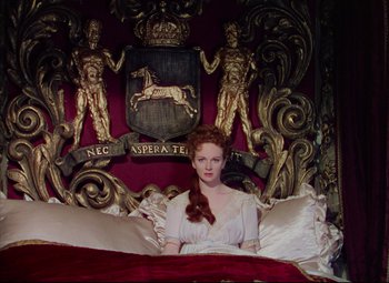Movie still from “Saraband” (1948), directed by Basil Dearden – A woman sitting on a bed in front of an ornate wall; Medium shot, Overhead angle