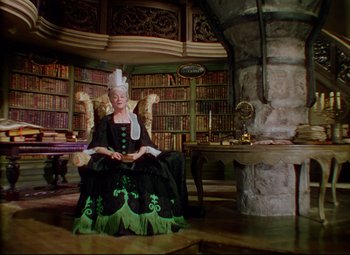 Movie still from “Saraband” (1948), directed by Basil Dearden – A woman dressed as a queen sitting in a chair in front of a bookcase filled with lots of books; Wide shot, High angle