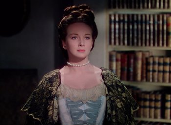 Movie still from “Saraband” (1948), directed by Basil Dearden – A woman wearing a dress and pearls in a room; Medium shot, Over the shoulder angle