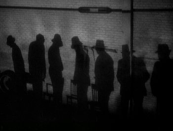 Movie still from “Scarface” (1932), directed by Richard Rosson – A black and white photo of a group of men standing next to each other in front of a brick wall; Wide shot, High angle
