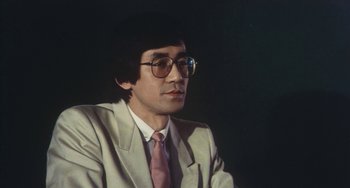 Movie still from “School in the Crosshairs” (1981), directed by Nobuhiko Ôbayashi – A man in a suit and tie sitting in front of a black background; Close Up shot, Low angle