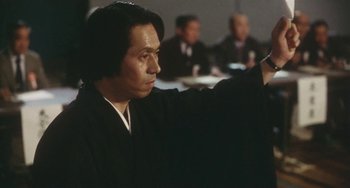 Movie still from “School in the Crosshairs” (1981), directed by Nobuhiko Ôbayashi – A man in a black robe is pointing at something in front of him; Close Up shot, Over the shoulder angle