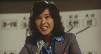 Movie still from “School in the Crosshairs” (1981), directed by Nobuhiko Ôbayashi – A woman in a gray jacket smiles while holding a microphone in her hand; Close Up shot, Low angle