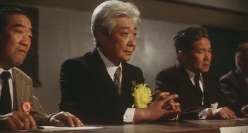 Movie still from “School in the Crosshairs” (1981), directed by Nobuhiko Ôbayashi – An older man wearing a suit sitting at a table; Medium shot, Low angle