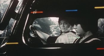 Movie still from “School in the Crosshairs” (1981), directed by Nobuhiko Ôbayashi – A man driving a car with another man in the reflection of the mirror; Close Up shot, Low angle