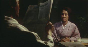 Movie still from “School in the Crosshairs” (1981), directed by Nobuhiko Ôbayashi – A woman sitting at a table reading a newspaper; Medium shot, Over the shoulder angle