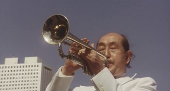 Movie still from “School in the Crosshairs” (1981), directed by Nobuhiko Ôbayashi – An older man playing a trumpet in front of a sky background; Extreme Close Up shot, Low angle