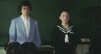 Movie still from “School in the Crosshairs” (1981), directed by Nobuhiko Ôbayashi – A man and a woman standing next to each other in front of a chalkboard; Medium shot, Over the shoulder angle