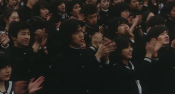 Movie still from “School in the Crosshairs” (1981), directed by Nobuhiko Ôbayashi – A group of people sitting and standing in front of each other clapping; Medium shot, High angle
