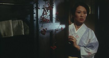 Movie still from “School in the Crosshairs” (1981), directed by Nobuhiko Ôbayashi – A woman standing in front of a window holding a cell phone; Medium shot, Over the shoulder angle