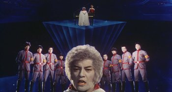 Movie still from “School in the Crosshairs” (1981), directed by Nobuhiko Ôbayashi – A group of people standing in front of an image of a man; Wide shot, Low angle