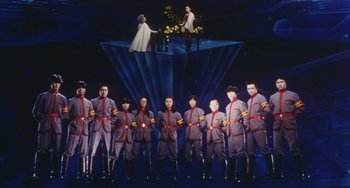 Movie still from “School in the Crosshairs” (1981), directed by Nobuhiko Ôbayashi – A group of men in grey uniforms standing in front of a stage; Wide shot, Low angle